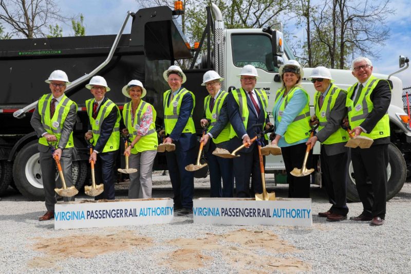 President Tim Sands with other officials wearing white hard hats and bright safety vests line up in front of a dump truck, each holding a gold shovel with fresh dirt for the ceremonial groundbreaking. 
