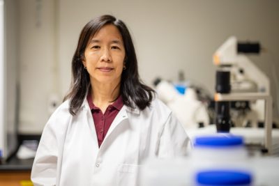 A woman in a white lab coat smiles at the camera in a lab full of equipment.