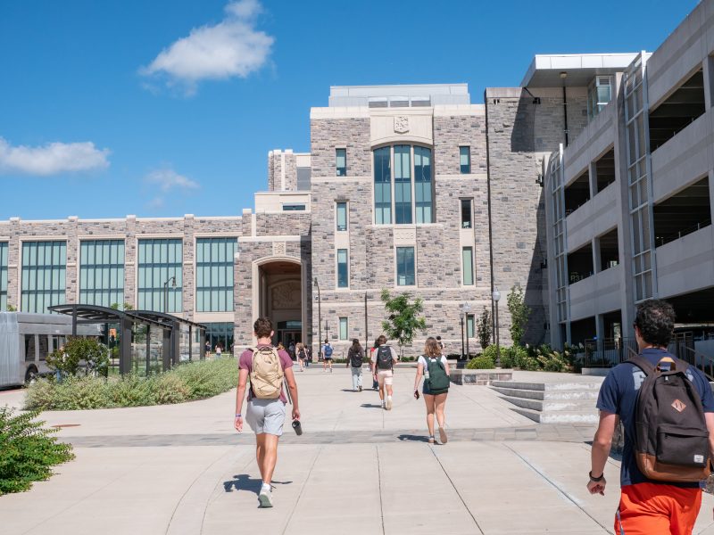 pedestrians walking on sidewalk under blue skies, bus stop and parking garage on either side of sidewalk